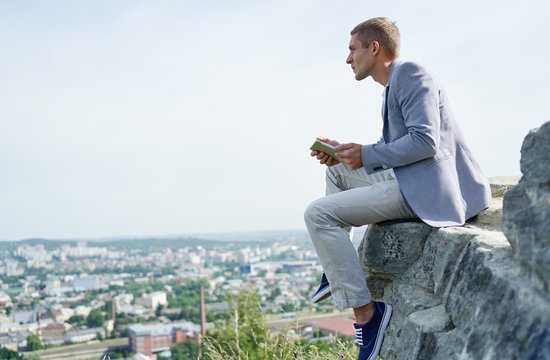 businessman with a tablet in his hands sits on a rock against the background of the city and looks into the distance