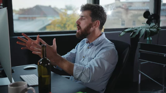 Fun drunk office executive working on computer doing stupid gestures. Overworked young business man falling asleep of alcohol intoxication.