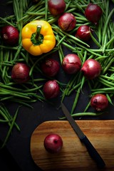 Green beans and red onions with green pepper scattered across a dark grey background. A wooden chopping board with a knife with a black handle sits next to a onion. Portrait flat lay or top view.
