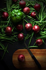 Green beans and red onions with green pepper scattered across a dark grey background. A wooden chopping board with a knife with a black handle sits next to a onion. Portrait flat lay or top view.