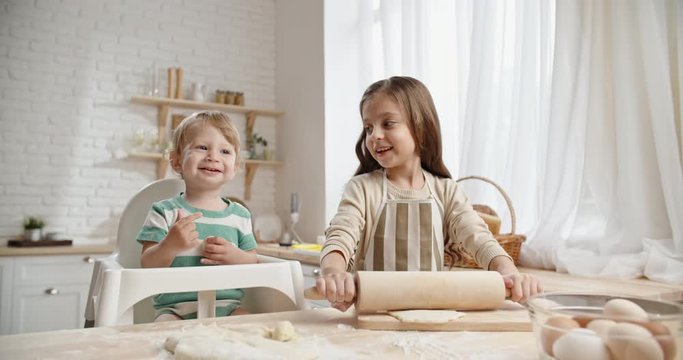 Little caucasian girl is learning how to bake, preparing pastry while her younger brother in baby chair is playing and positively smiling - togetherness, childhood dream concept 4k