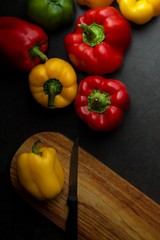 Red, yellow and green peppers against a dark grey background.  A portrait flat lay view of an assortment of peppers.   Chopping board with knife with black handle. Ideal dark background with space for