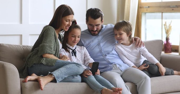 Little schoolgirl relaxing on sofa with smiling parents and younger brother, showing funny video on smartphone. Full length happy family using mobile applications, enjoying leisure time together.