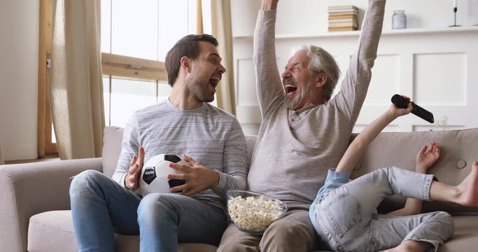 Happy middle aged man enjoying watching sport championship on smart tv with young son and small kid grandson at home. Overjoyed family supporting soccer team, celebrating win victory together.