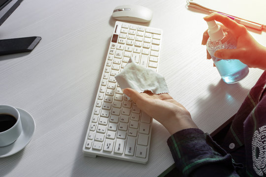A girl in a checked shirt splashes cleaning agent on a rag to clean the keyboard. A Cup of coffee, a mouse, and a Notepad with a pen on a white office Desk. The concept of cleaning of the office