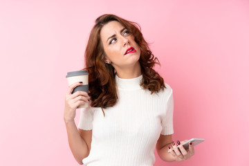 Young Russian woman over isolated pink background holding coffee to take away and a mobile while thinking something