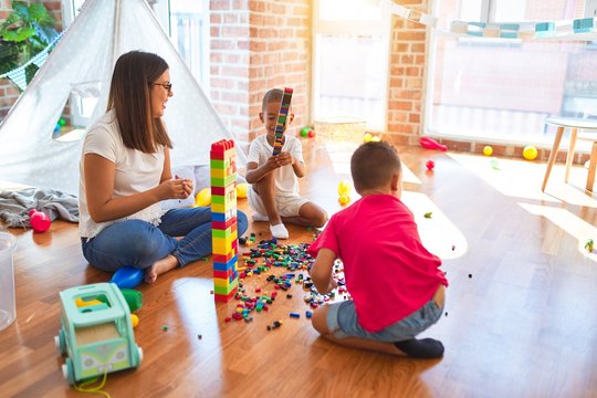 Young beautiful teacher and toddlers playing with building blocks around lots of toys at kindergarten