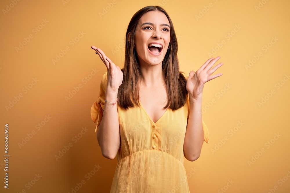 Wall mural young beautiful brunette woman wearing casual dress standing over yellow background celebrating mad 