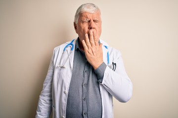 Senior handsome hoary doctor man wearing coat and stethoscope over white background bored yawning tired covering mouth with hand. Restless and sleepiness.