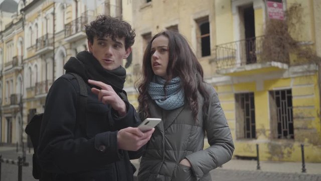 Portrait of young Caucasian couple of travelers using online map in smartphone. Lost disoriented boy and girl standing in foreign city and looking around. Tourism, travelling, lifestyle, journey.