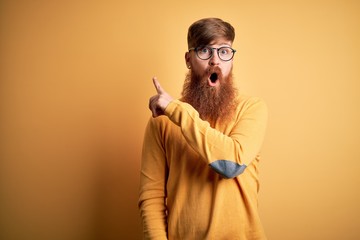 Handsome Irish redhead man with beard wearing glasses over yellow isolated background Surprised pointing with finger to the side, open mouth amazed expression.