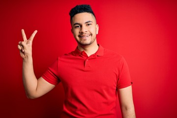 Fototapeta premium Young handsome man wearing red casual polo standing over isolated background showing and pointing up with fingers number two while smiling confident and happy.