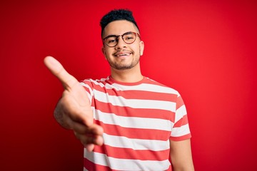 Young handsome man wearing casual striped t-shirt and glasses over isolated red background smiling cheerful offering palm hand giving assistance and acceptance.