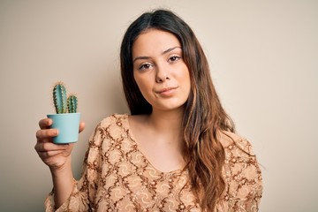 Young beautiful brunette woman holding cactus pot over isolated white background with a confident expression on smart face thinking serious