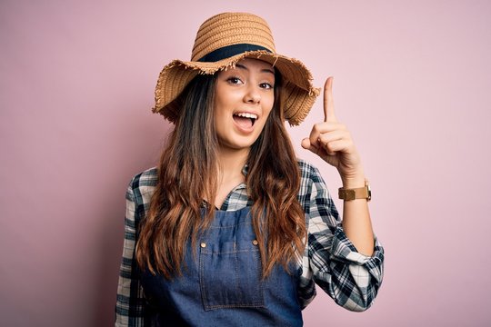 Young beautiful brunette farmer woman wearing apron and hat over pink background pointing finger up with successful idea. Exited and happy. Number one.