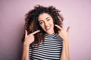 Young beautiful woman with curly hair and piercing wearing casual striped t-shirt smiling cheerful showing and pointing with fingers teeth and mouth. Dental health concept.