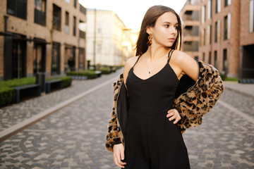 Fototapeta premium Gorgeous girl walks about the city. Brunette with long hair in a black dress and a leopard cape. Girl, fashion photo, street fashion. Fashionable girl walks in the city of Europe, travels.