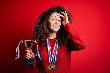 Young beautiful successful woman with curly hair and piercing holding trophy wearing medals stressed with hand on head, shocked with shame and surprise face, angry and frustrated. Fear and upset for 