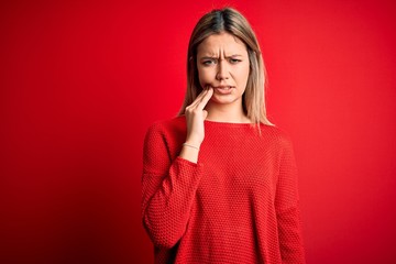 Young beautiful blonde woman wearing casual sweater over red isolated background touching mouth with hand with painful expression because of toothache or dental illness on teeth. Dentist