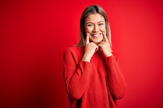 Young beautiful blonde woman wearing casual sweater over red isolated background Smiling with open mouth, fingers pointing and forcing cheerful smile
