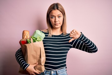 Young beautiful woman holding paper bag with purchase over isolated pink background with angry...