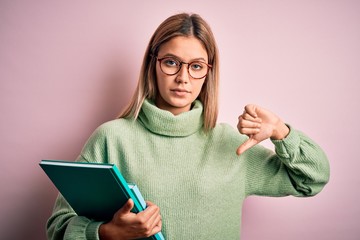 Young beautiful woman wearing glasses holding books over isolated pink background with angry face,...