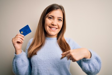 Young blonde woman holding credit card as payment over isolated background with surprise face pointing finger to himself