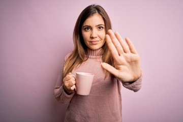 Young blonde woman drinking a cup of coffee over pink isolated background with open hand doing stop sign with serious and confident expression, defense gesture