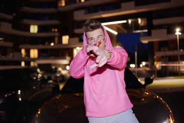 A man with glasses at night. Fashion guy photo nightly. The guy has fun at night on the street. Rep men. Teen at night near a golden car in a pink sweater. Photo of a fashionable guy who is crazy.