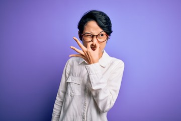 Young beautiful asian girl wearing casual shirt and glasses standing over purple background smelling something stinky and disgusting, intolerable smell, holding breath with fingers on nose. Bad smell