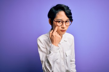 Young beautiful asian girl wearing casual shirt and glasses standing over purple background Pointing to the eye watching you gesture, suspicious expression
