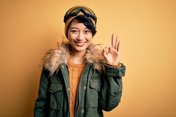 Beautiful asian skier girl wearing snow sportswear using ski goggles over yellow background smiling positive doing ok sign with hand and fingers. Successful expression.