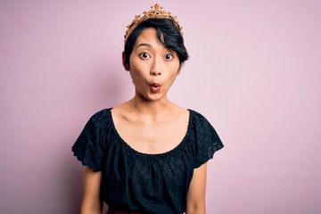 Young beautiful chinese woman wearing golden crown of king over isolated pink background afraid and shocked with surprise expression, fear and excited face.