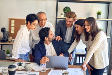 Group of business workers smiling happy and confident. One of them sitting and partners standing around. Working together with smile on face looking at the laptop at the office