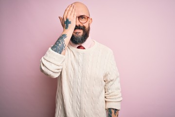 Handsome bald man with beard and tattoo wearing glasses and sweater over pink background covering one eye with hand, confident smile on face and surprise emotion.