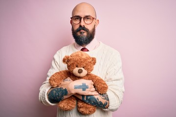 Handsome bald man with beard and tattoo holding teddy bear over isolated pink background with a...