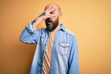 Handsome bald man with beard wearing casual denim jacket and striped t-shirt peeking in shock covering face and eyes with hand, looking through fingers with embarrassed expression.