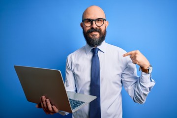 Handsome bald business man with beard working using laptop over blue background with surprise face pointing finger to himself