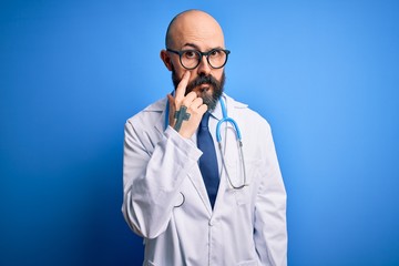 Handsome bald doctor man with beard wearing glasses and stethoscope over blue background Pointing to the eye watching you gesture, suspicious expression