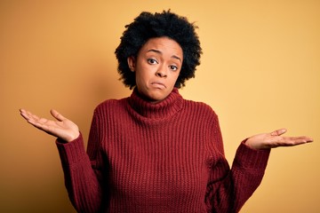 Young beautiful African American afro woman with curly hair wearing casual turtleneck sweater clueless and confused expression with arms and hands raised. Doubt concept.