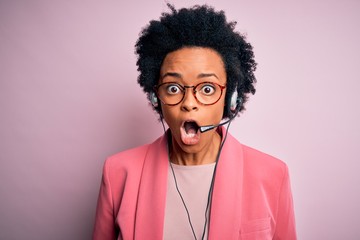 Young African American call center operator woman with curly hair using headset afraid and shocked...