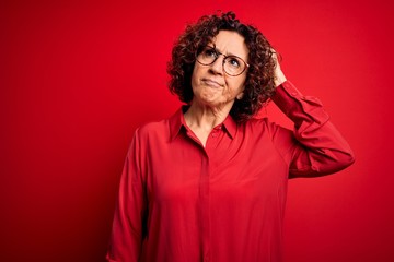 Middle age beautiful curly hair woman wearing casual shirt and glasses over red background confuse and wondering about question. Uncertain with doubt, thinking with hand on head. Pensive concept.