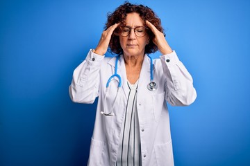 Middle age curly hair doctor woman wearing coat and stethoscope over blue background with hand on head for pain in head because stress. Suffering migraine.