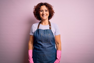 Middle age curly hair woman cleaning doing housework wearing apron and gloves with a happy and cool smile on face. Lucky person.