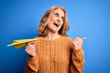 Middle age beautiful blonde woman holding paper airplane over isolated blue background pointing and showing with thumb up to the side with happy face smiling