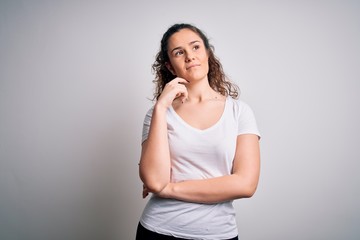 Young beautiful woman with curly hair wearing casual t-shirt standing over white background with hand on chin thinking about question, pensive expression. Smiling with thoughtful face. Doubt concept.