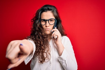 Young beautiful woman with curly hair having conversation talking on the smartphone pointing with finger to the camera and to you, hand sign, positive and confident gesture from the front