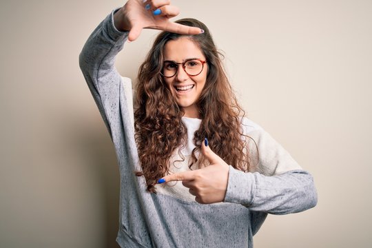 Young beautiful woman with curly hair wearing sweater and glasses over white background smiling making frame with hands and fingers with happy face. Creativity and photography concept.