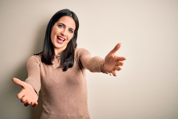 Young brunette woman with blue eyes wearing casual sweater over isolated white background smiling cheerful offering hands giving assistance and acceptance.