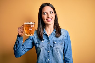 Young woman with blue eyes drinking jar of beer standing over isolated yellow background smiling...